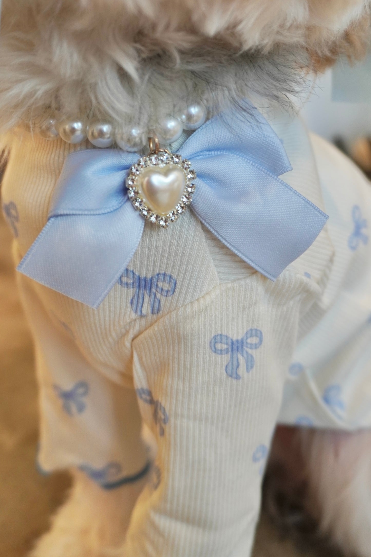 Close-up of Heartfelt Pearl Dog Necklace featuring a pearl strand, blue bow, and rhinestone heart charm on cream and blue ruffle dog top