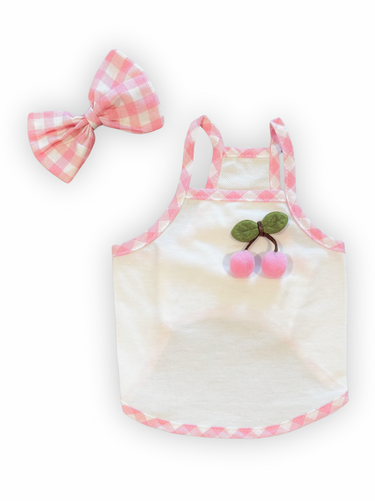 Small dog wearing sleeveless cherry print top with pink gingham trim and matching clip-on bow