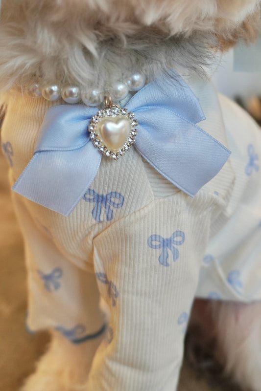 Close-up of Heartfelt Pearl Dog Necklace featuring a pearl strand, blue bow, and rhinestone heart charm on cream and blue ruffle dog top