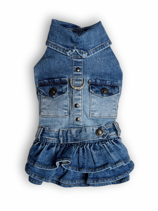 A blue denim dog dress with a ruffled layered skirt, collar neck, and sleeveless design. It includes two non-functional pockets and a D-ring for leash attachment.